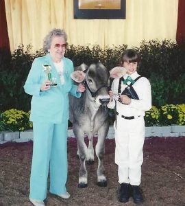 The dairy judge is wearing a blue pantsuit holding a trophy next to a Brown Swiss heifer. Hannah is standing next to the heifer after showing it at a 4-H show. 