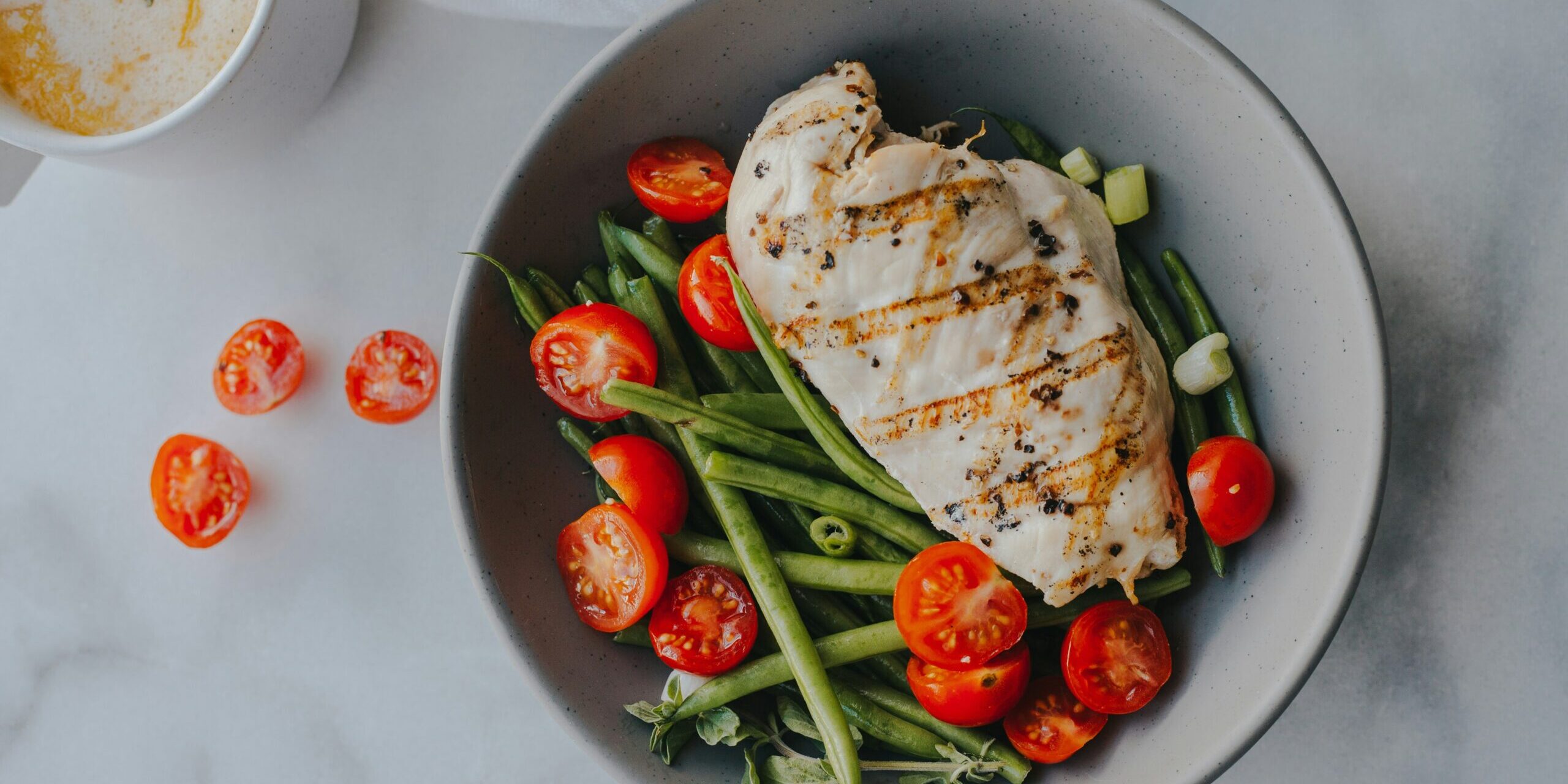 A cooked slice of chicken breast in a bowl of green beans and tomatoes on a white counter top
