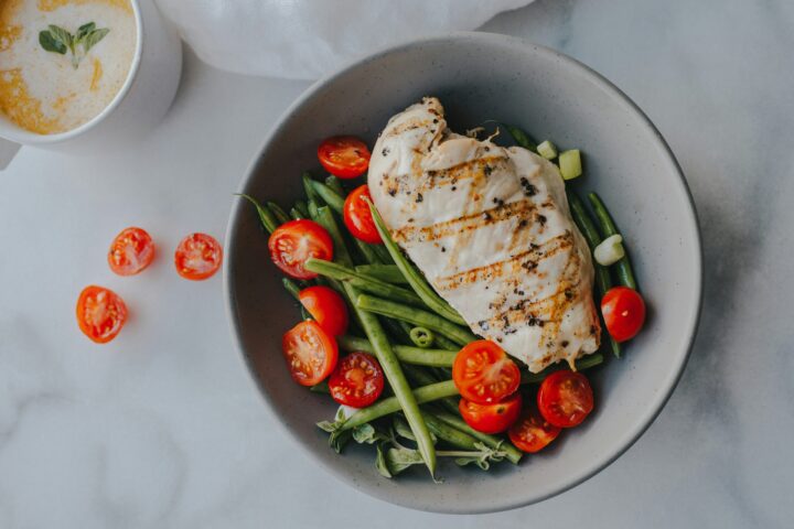 A cooked slice of chicken breast in a bowl of green beans and tomatoes on a white counter top
